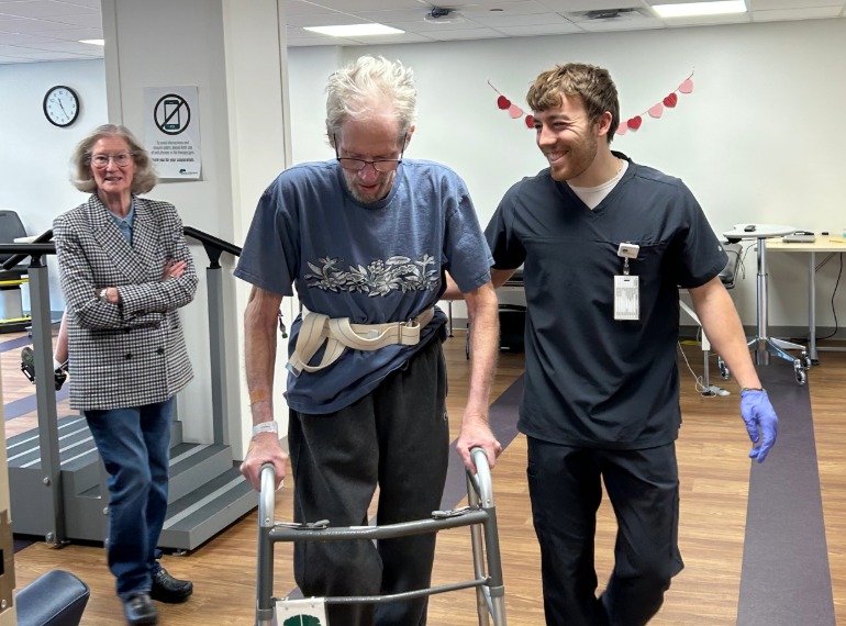 Jeff walking with a walker with a therapist and his wife.