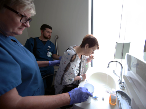 A patient leans over the sink to brush her teeth with the help or her care team to remain stable.