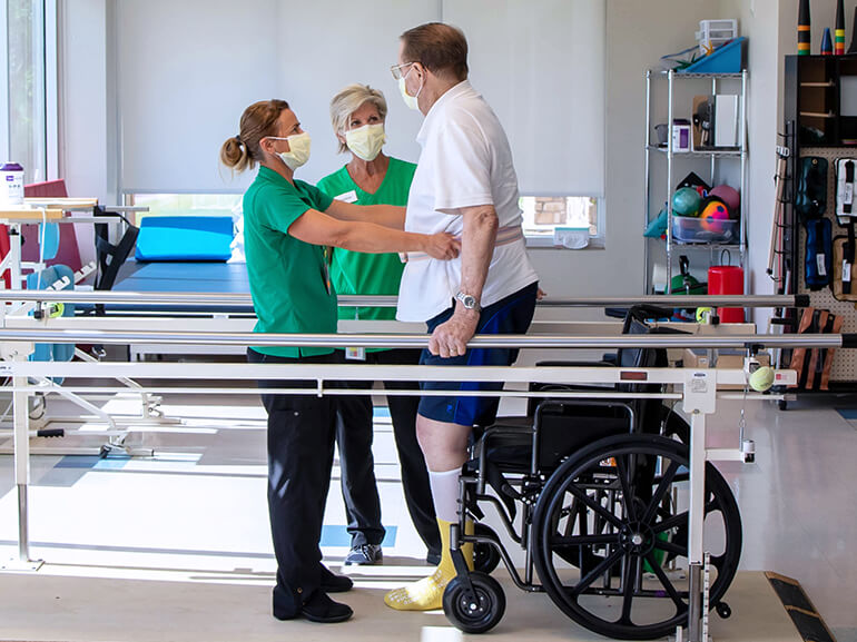 Two physical therapists assisting a patient on the parallel bars