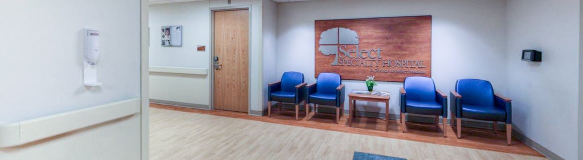A view of Select Specialty Hospital Downriver upon entering featuring a few blue chairs and a sign with the hospital name.
