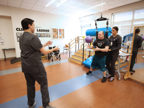 A patient stands with the help of an overhead support system. He plays catch with her therapist while working on balance.