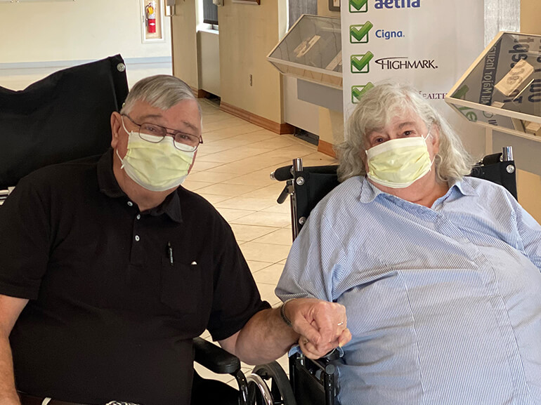 Art and Nancy wearing Covid masks and holding hands in their wheelchairs in hospital lobby.