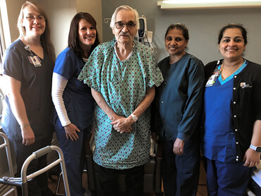 John standing on his own with his care team around him in his hospital room.