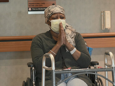 Matilda wearing a mask and sitting in a wheelchair with hands clasped in prayer.