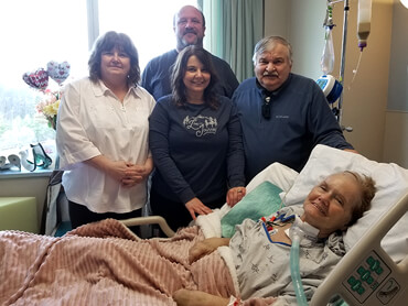 Patsy using airway support and smiling from her hospital bed with family standing behind her.