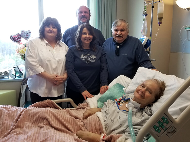 Patsy using airway support and smiling from her hospital bed with family standing behind her.
