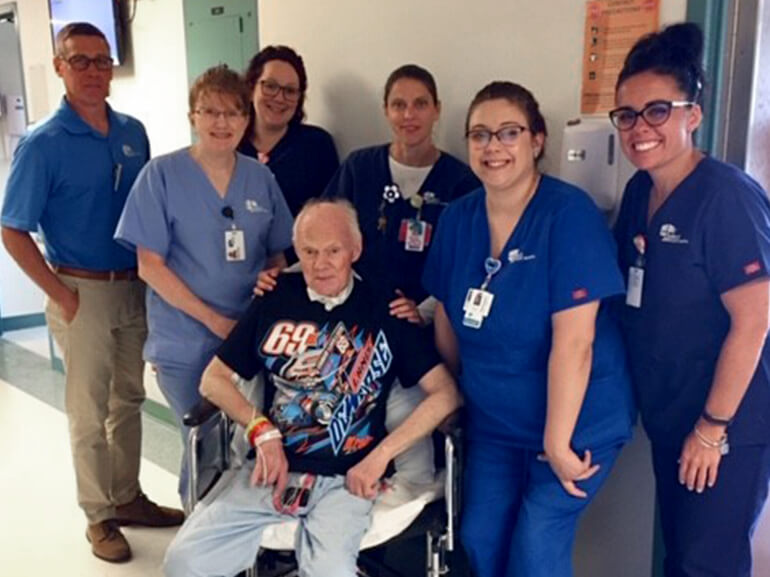 Ray, wearing a t-shirt of his favorite stock car driver Lance Dewease, sits in a chair surrounded by his care team.