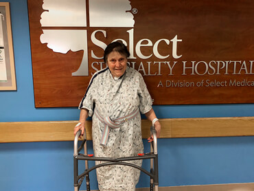 Rosemary stands in the hallway with the support of a walker in front of Select Specialty Hospital sign.