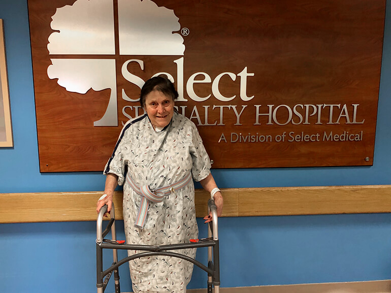 Rosemary smiles and stands with her walker in the hospital hallway.