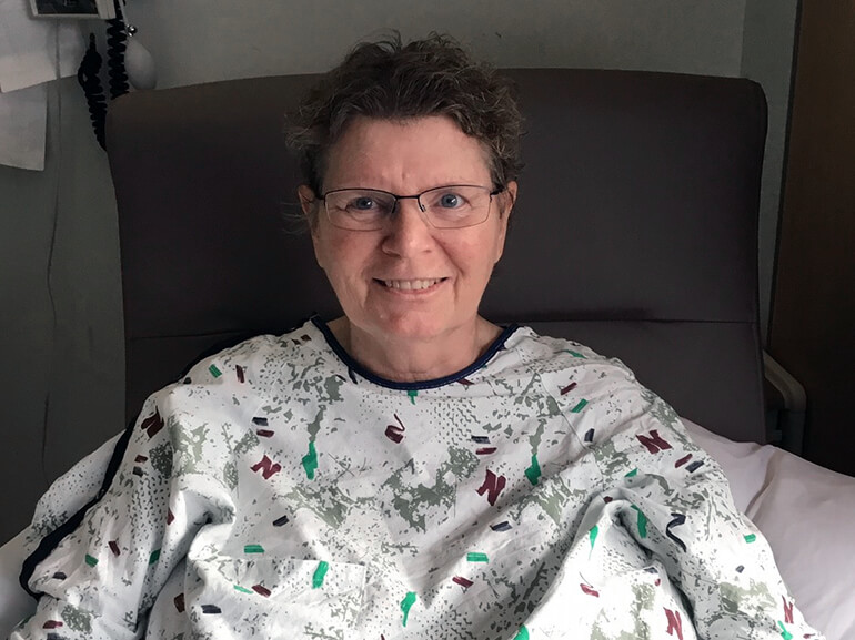 Vicki wearing a medical gown and sitting upright in a chair in her hospital room.