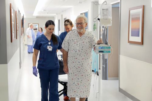 A patient holds his IV pole while walking down the hospital hallway with his care team's support.