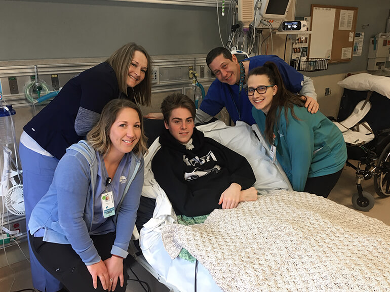 Sam lies in a hospital bed while five caregivers stand around the bed in a hospital room with medical equipment visible.
