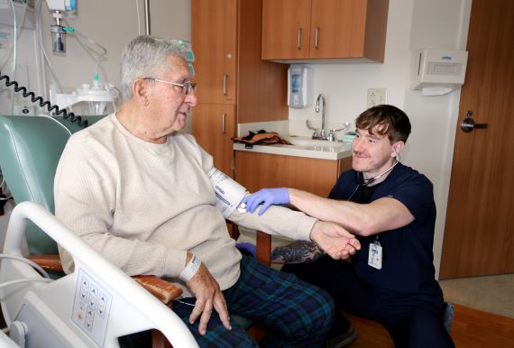 A nurse crouches down to take a patients blood pressure as they sit in a chair beside the hospital bed.