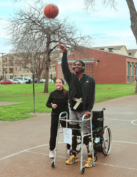 Patient is shooting a basketball at a hoop with the support of a walker and occupational therapist beside him