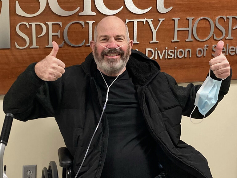 Keith smiling in his wheelchair with two thumbs up.
