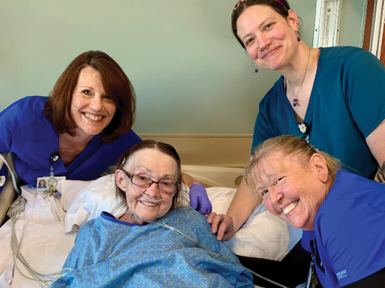 Sharon Seidl, lying in a hospital bed, surrounded by three female nurses, all smiling.