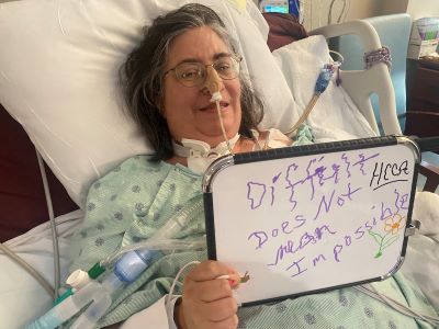 Stacy holds up a whiteboard while laying in her hospital bed. The whiteboard reads, "difficult does not mean impossible". 