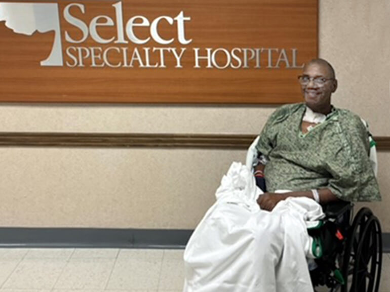 Stephen Sims sits in his wheelchair in front of the Select Specialty Hospital sign, and smiles for the camera.